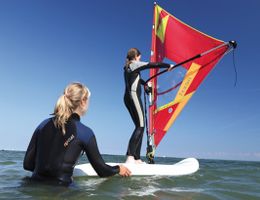 Zwei Personen beim Windsurftraining im Meer, eine steht auf dem Surfbrett mit Segel, die andere hält das Brett im Wasser.